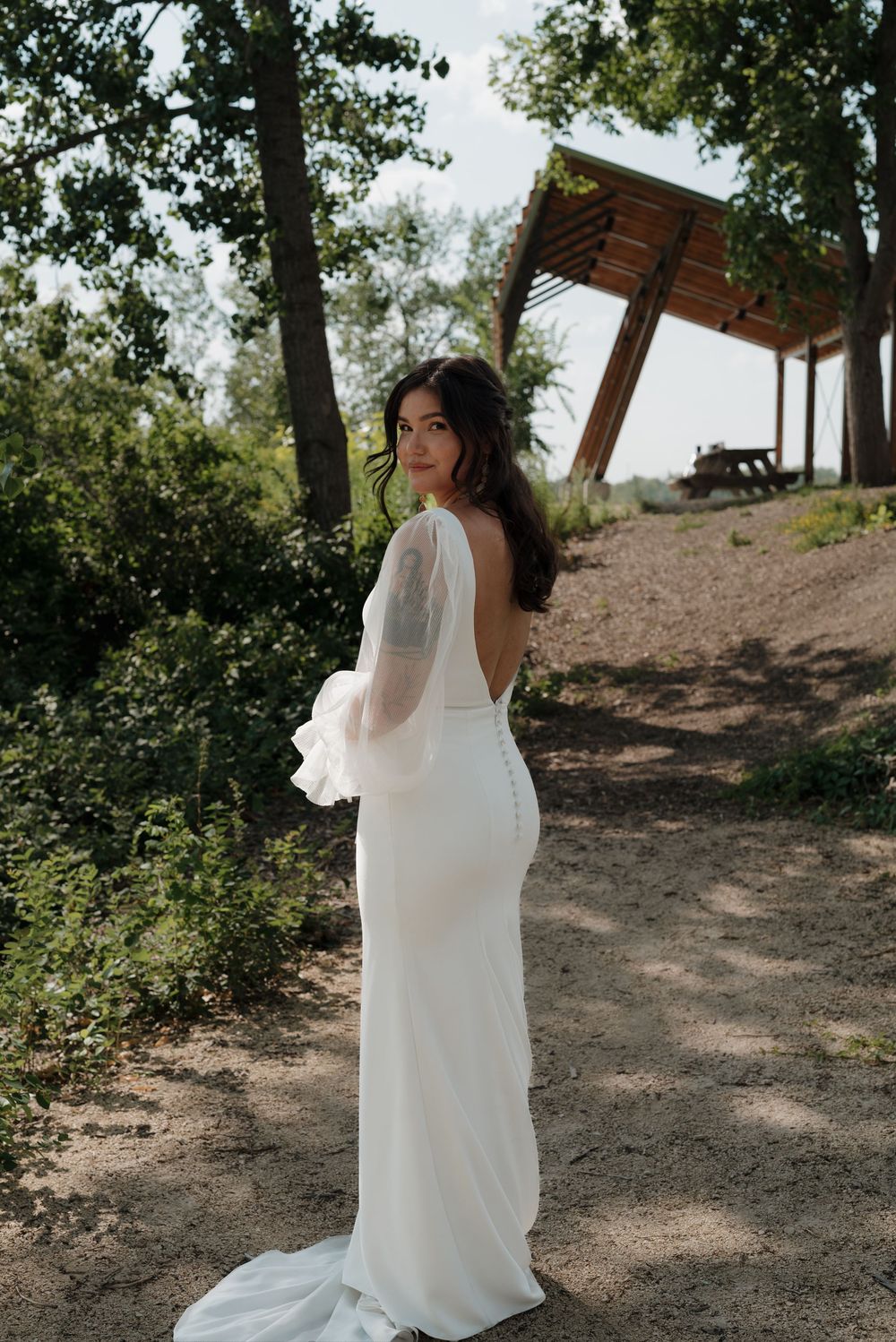 A person in a white wedding dress with sheer sleeves stands on a dirt path under a wooden shelter in nature.