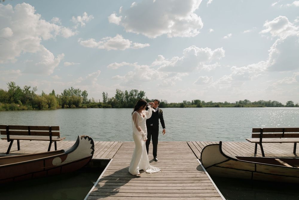 Two people in formal attire stand together on a lakeside dock with benches under a bright cloudy sky.