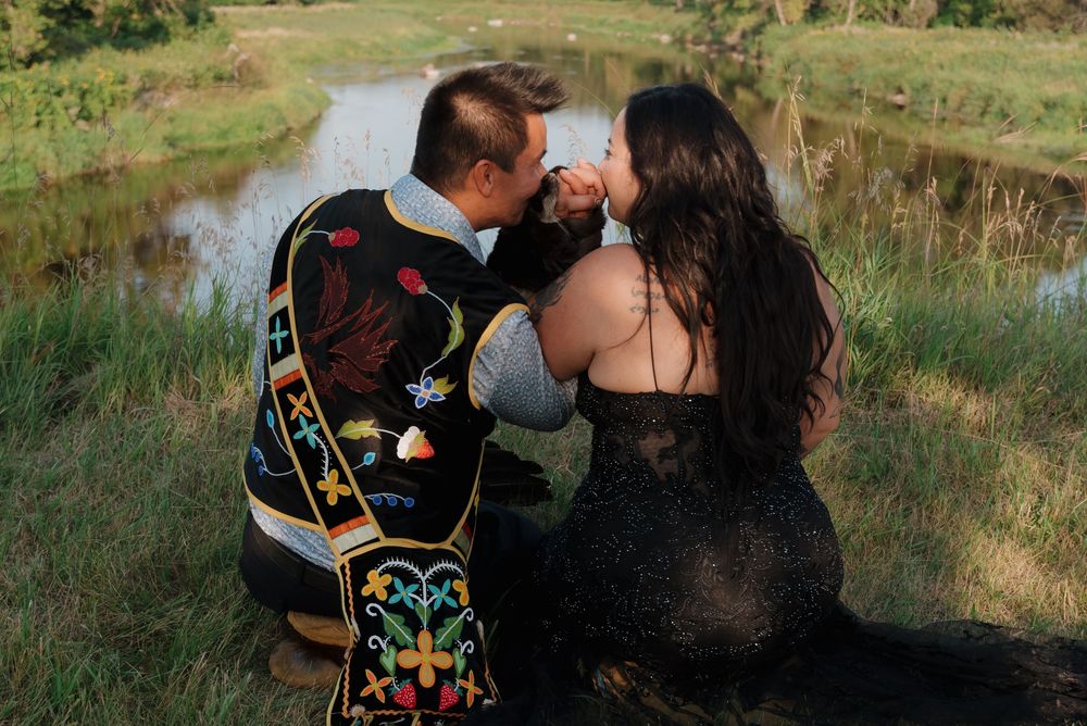 Two people in elegant attire share an intimate moment near a tranquil pond surrounded by green grass and nature.