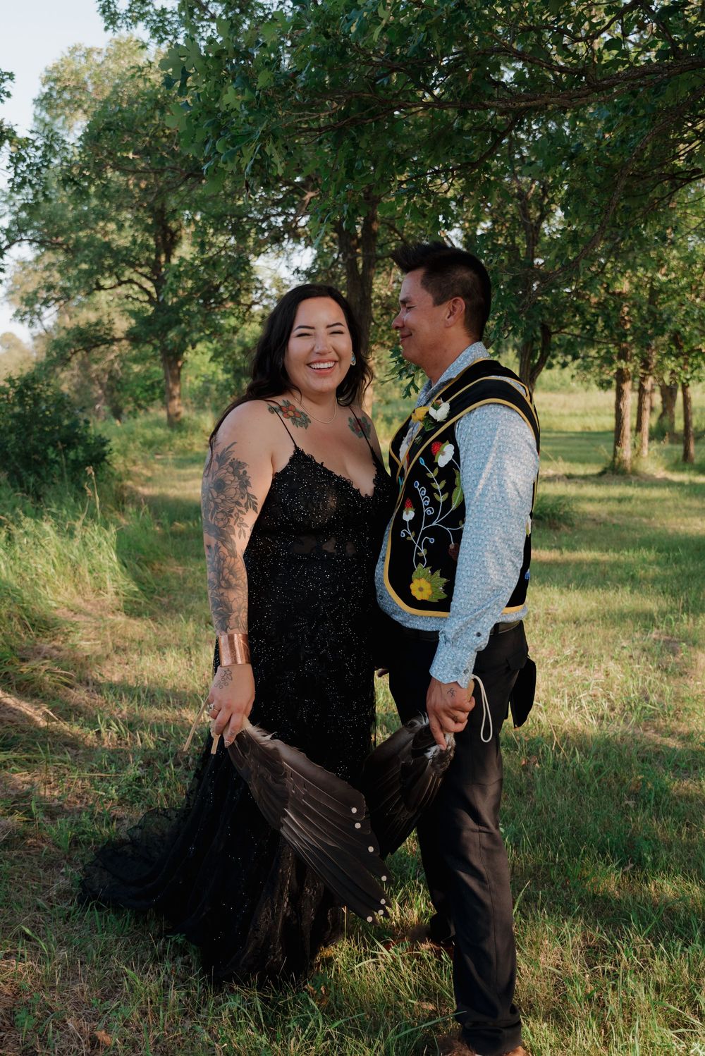 A romantic couple poses outdoors in formal attire, with a black dress and traditional regalia against a green nature backdrop.