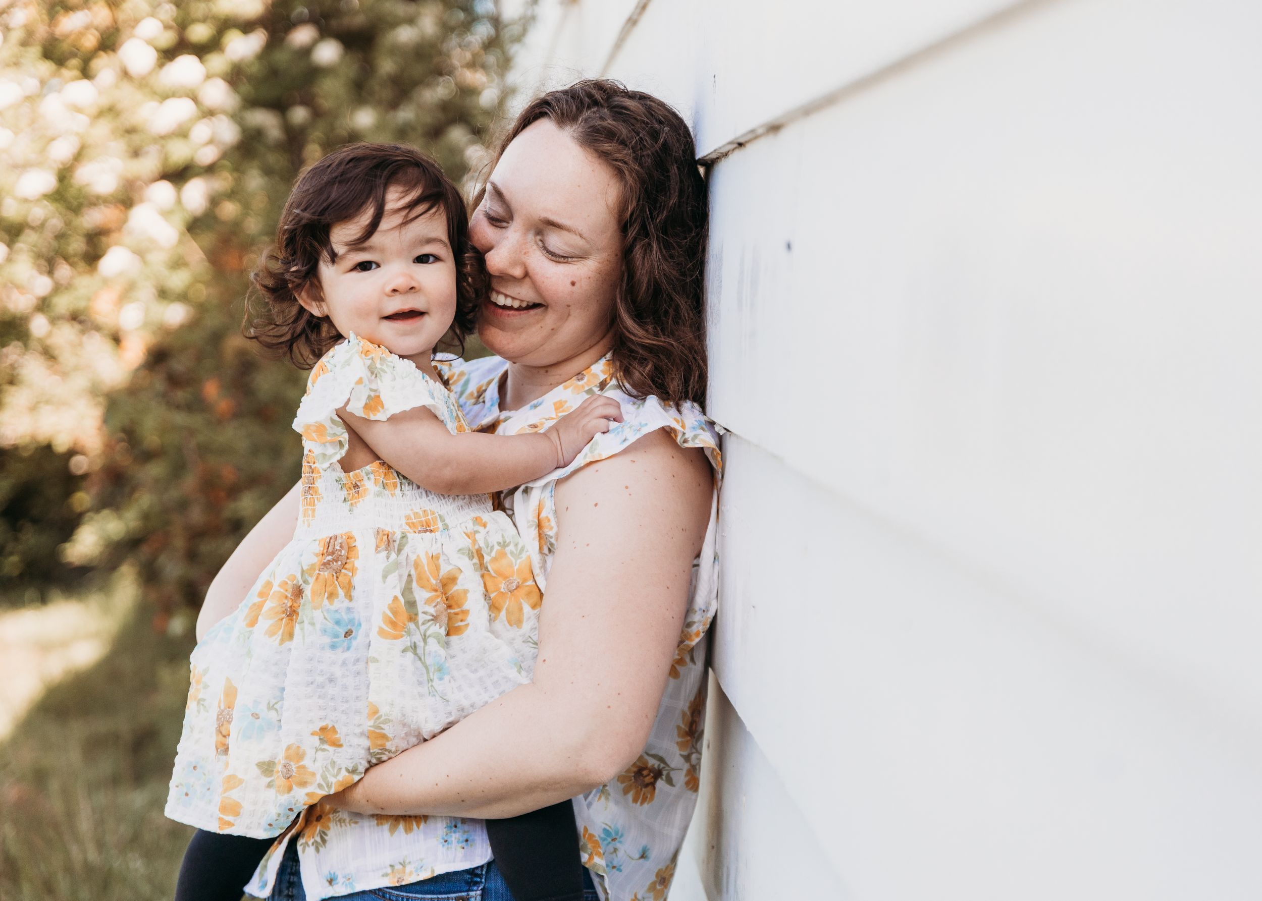 Parent and toddler share tender moments in a series of outdoor portraits against a white wall in golden sunlight.