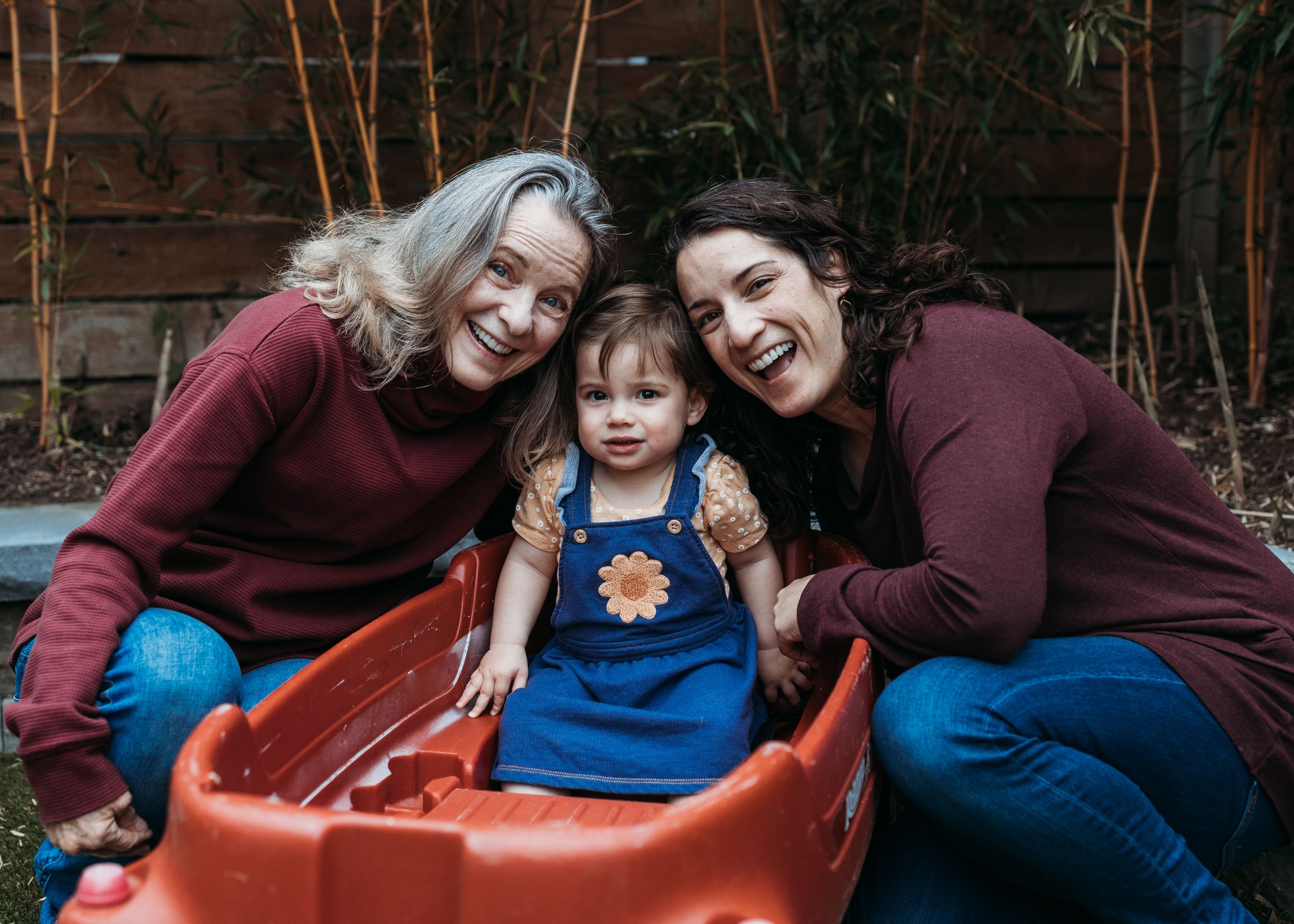 Three generations gather around a red toy wagon in a backyard setting wearing burgundy sweaters and denim.