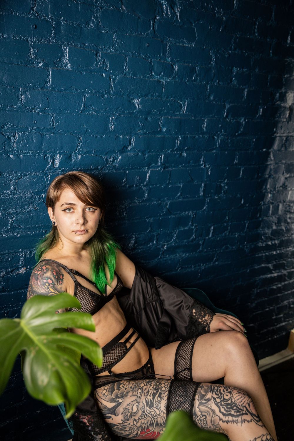 Model poses by monstera plant against dark blue brick wall.