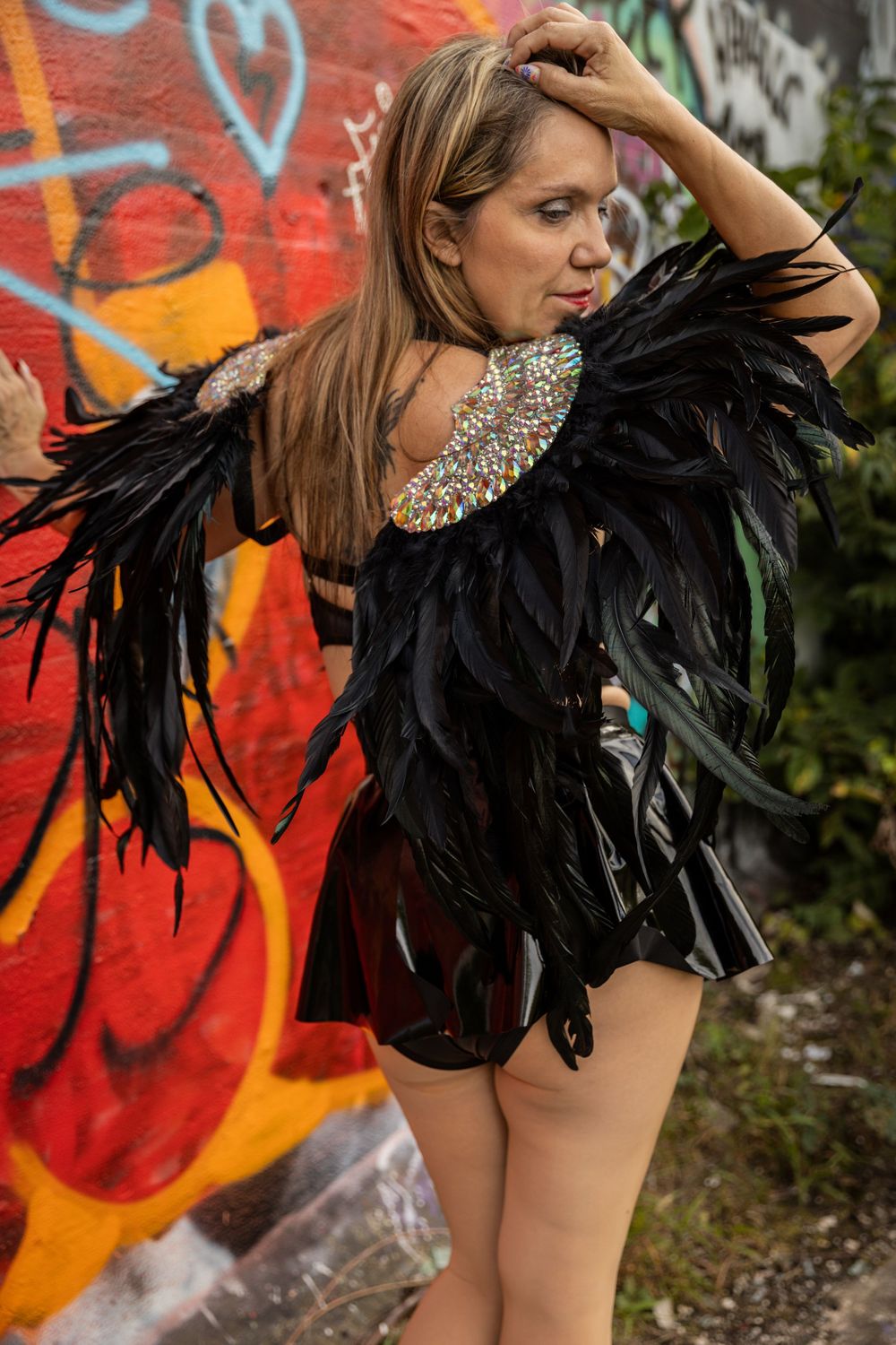 Artistic fashion photography featuring black feather costume details against urban graffiti background.