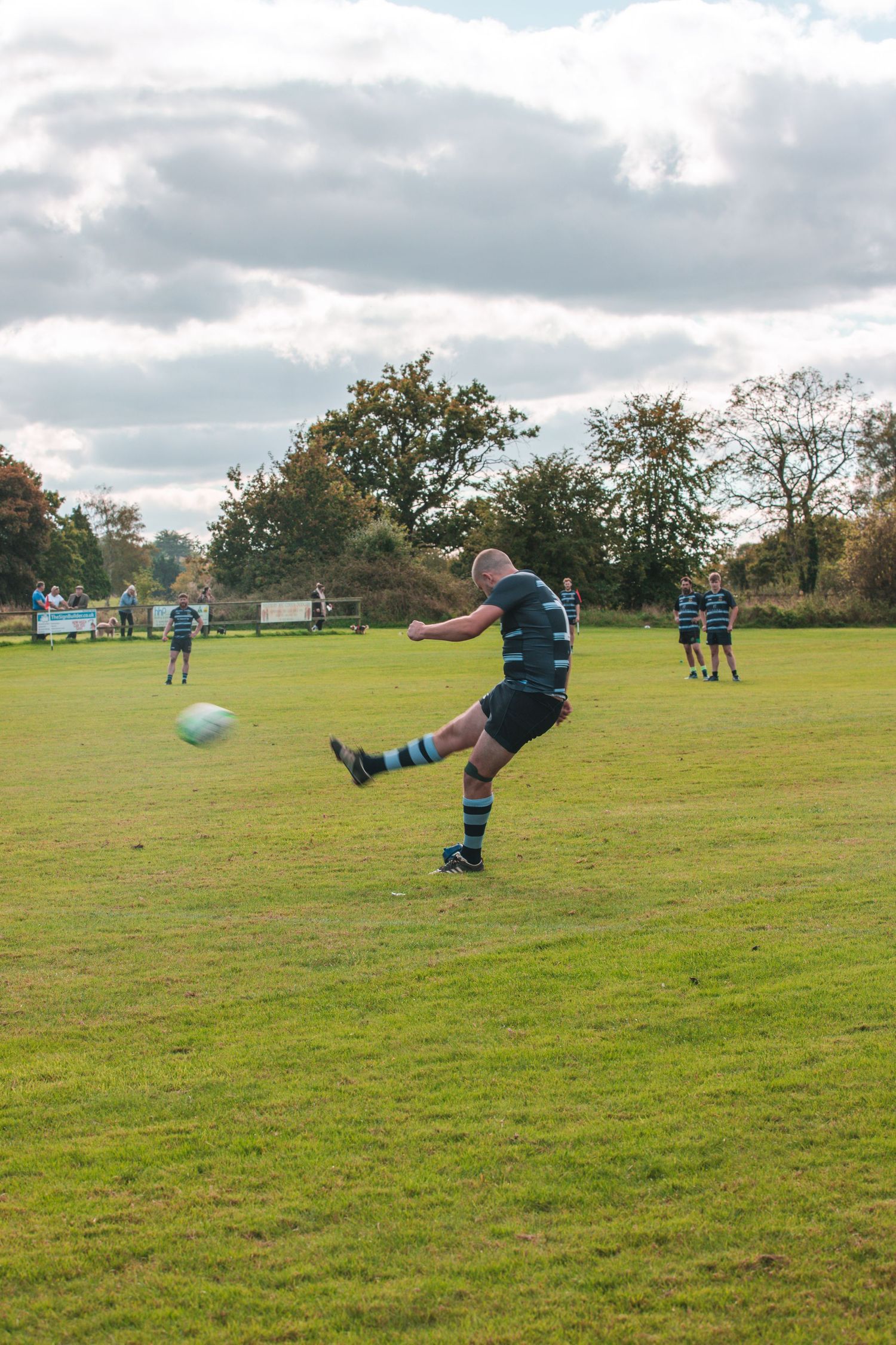 Rugby player executing a kicking motion on a green field with cloudy skies overhead.
