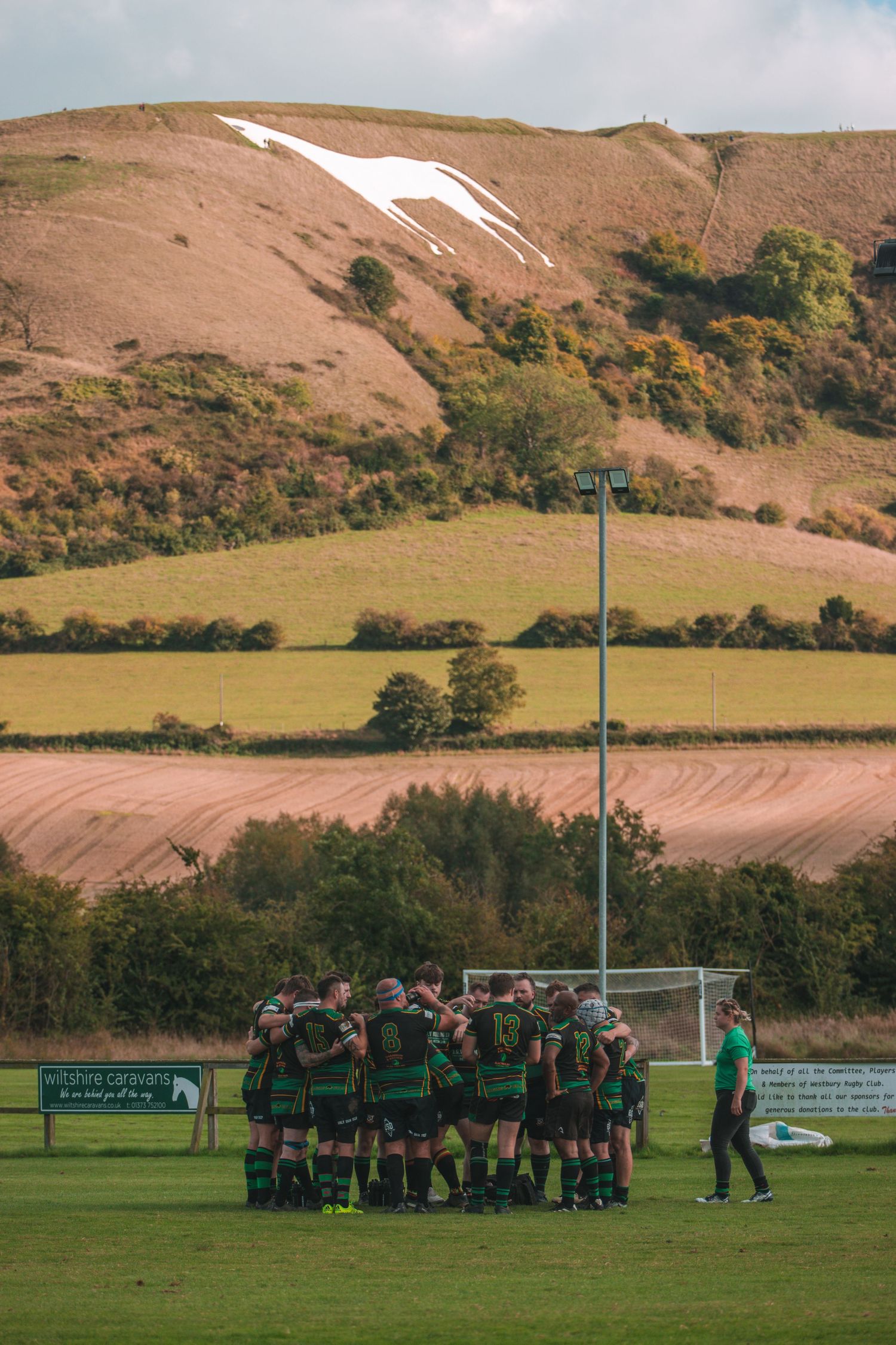 Rugby team huddles on field with iconic white horse chalk figure visible on distant hillside.