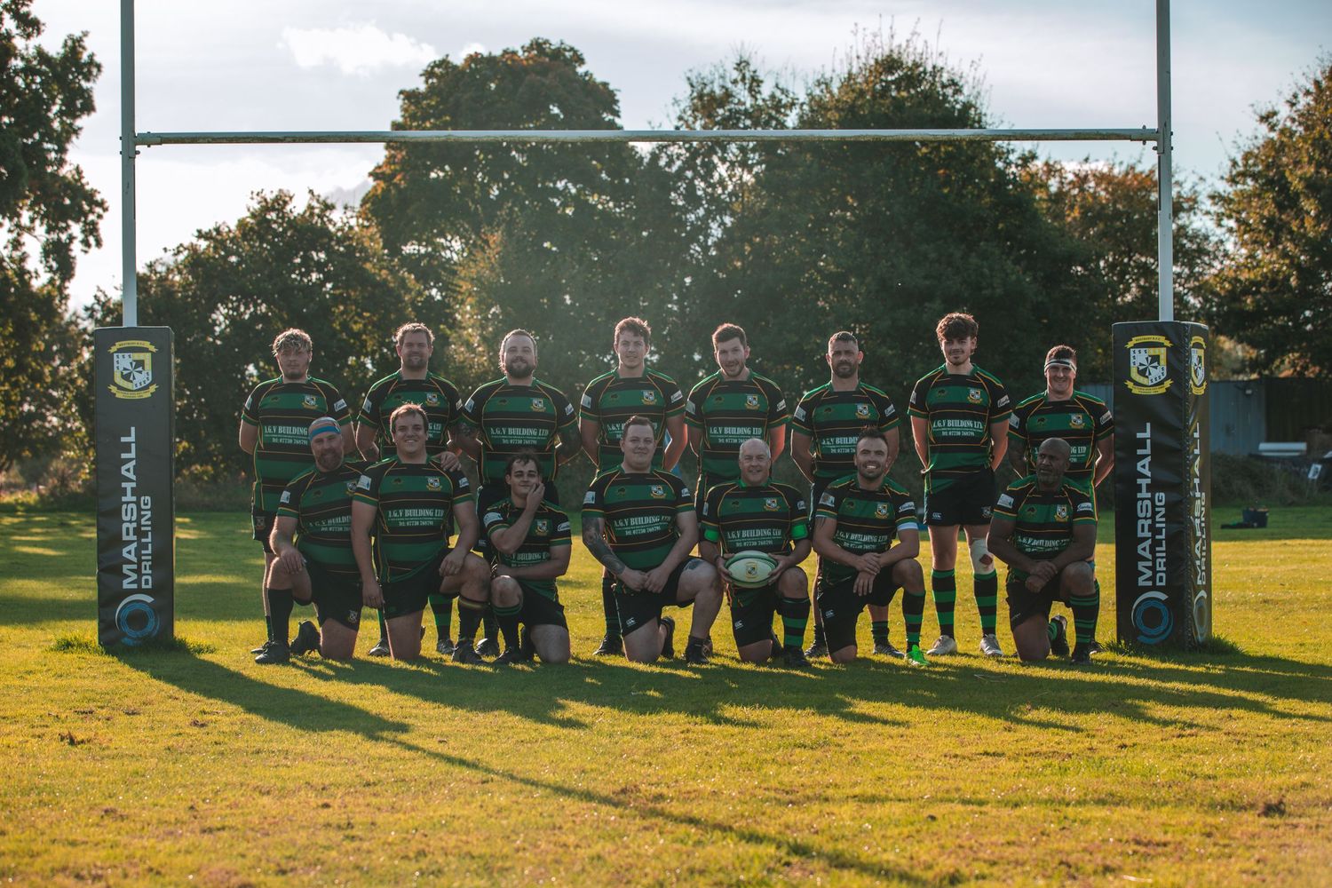 Rugby team in green and black striped jerseys poses under goal posts for group photo.