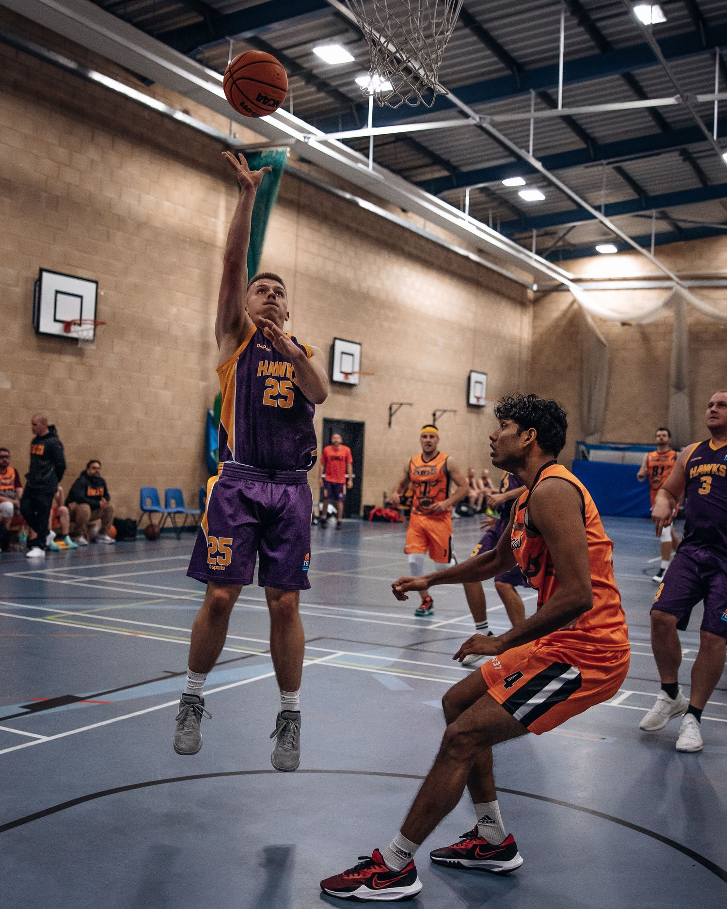 Indoor basketball game action with players in purple and orange jerseys competing for a shot.