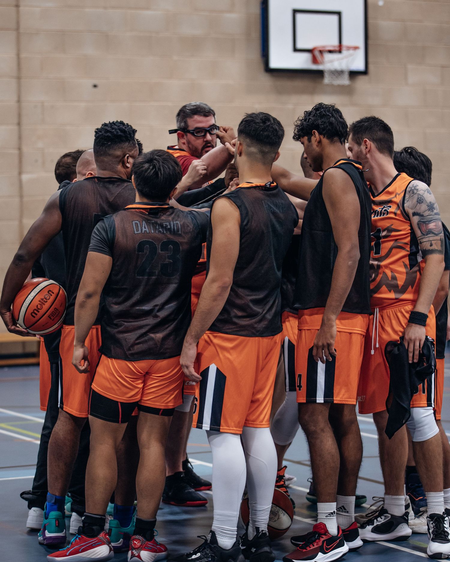 Basketball team huddles together during game discussion wearing orange and black uniforms.