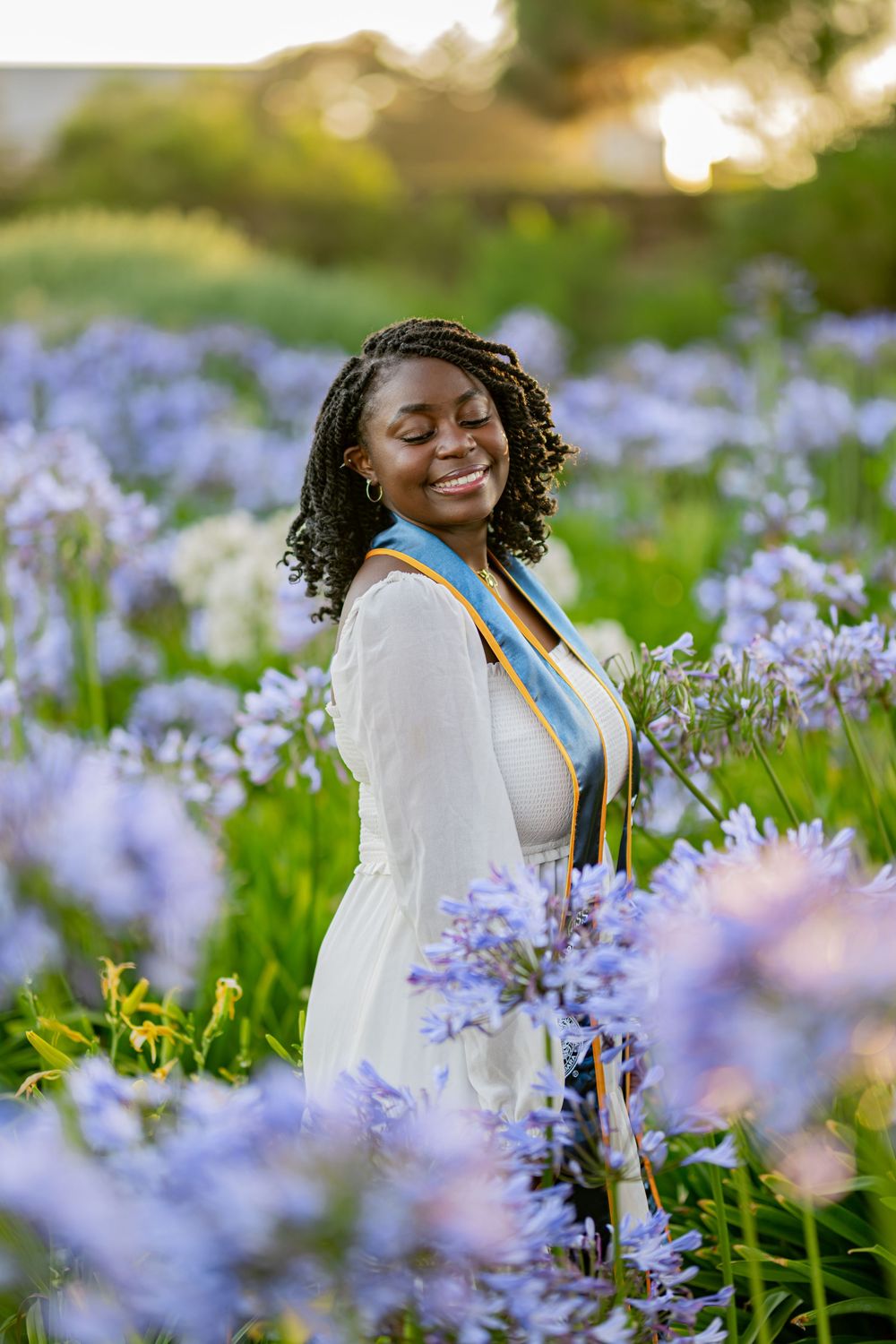 A person in a white dress sits joyfully among blooming purple wildflowers in a sunny meadow.