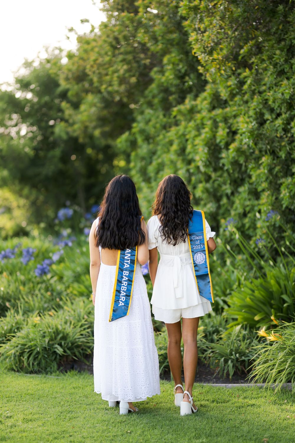 Two people in white dresses walk through a lush garden with blue ribbons against a backdrop of green trees and flowers.