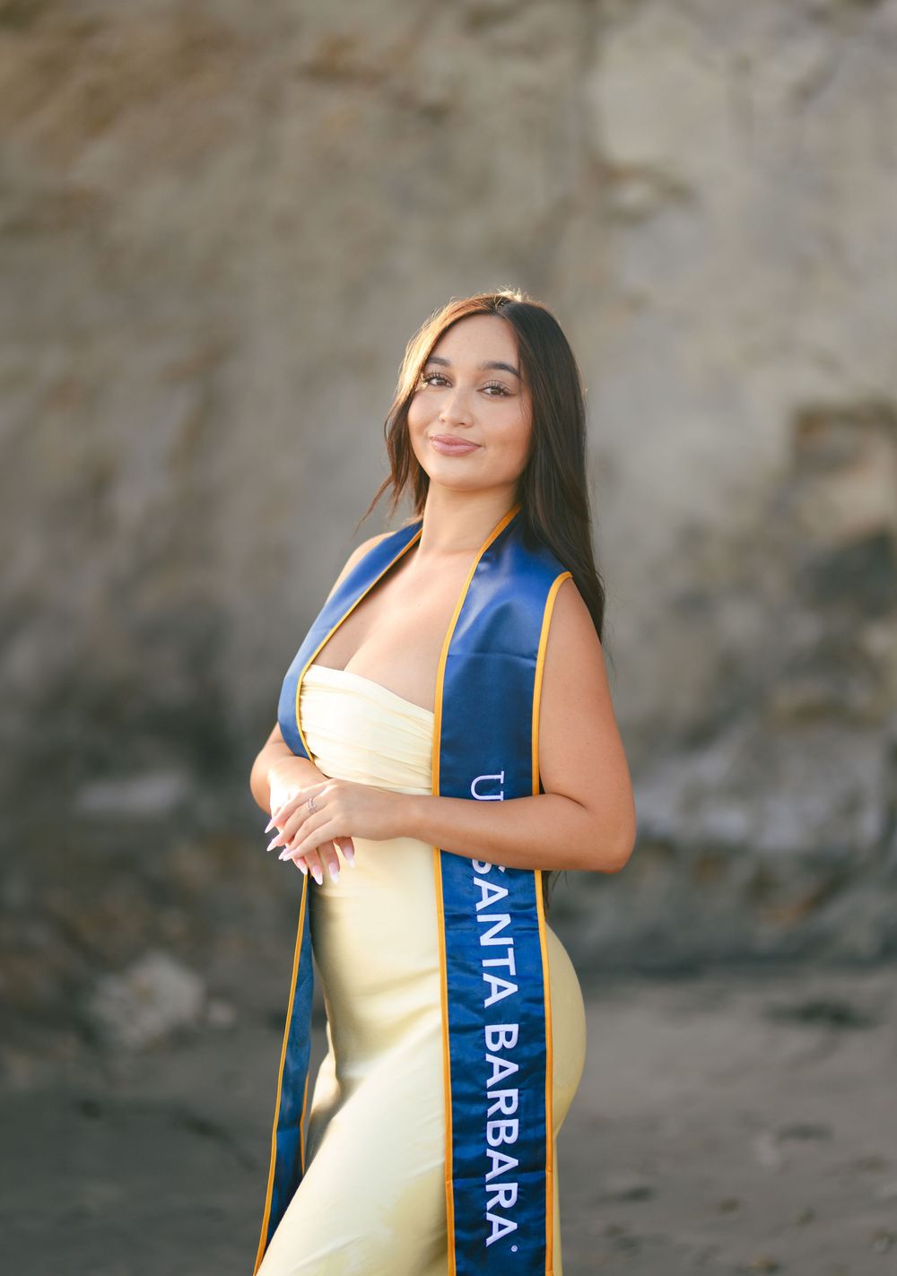 Young woman in yellow gown wearing UC Santa Barbara sash poses outdoors at rocky beach location.