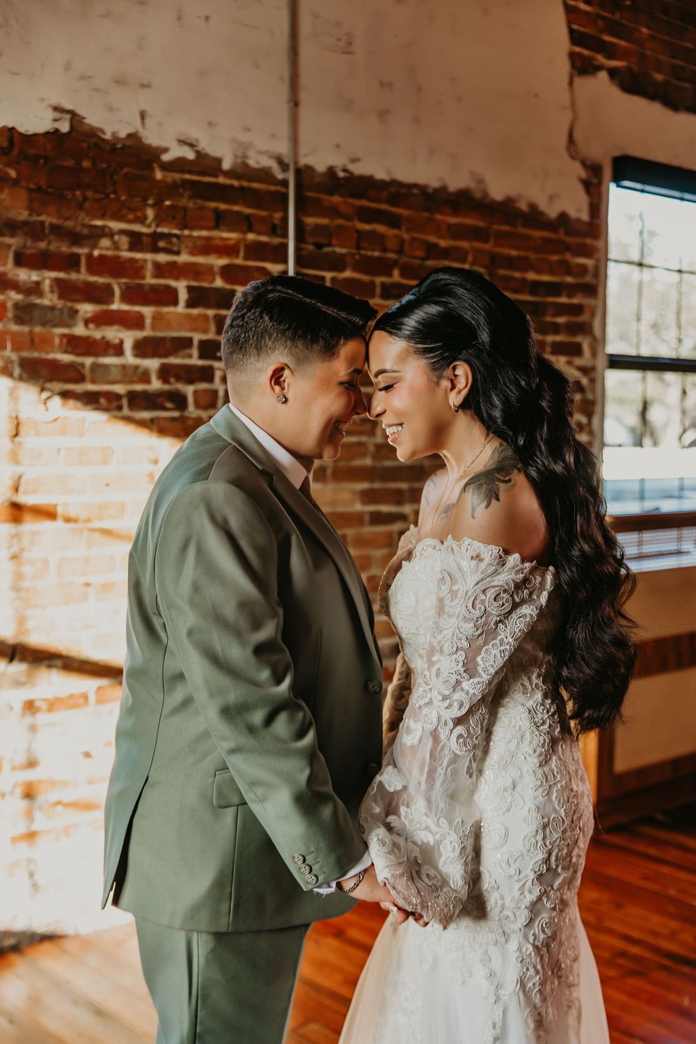 A couple in wedding attire holds hands and touches foreheads tenderly in a rustic brick-walled venue with warm sunlight.