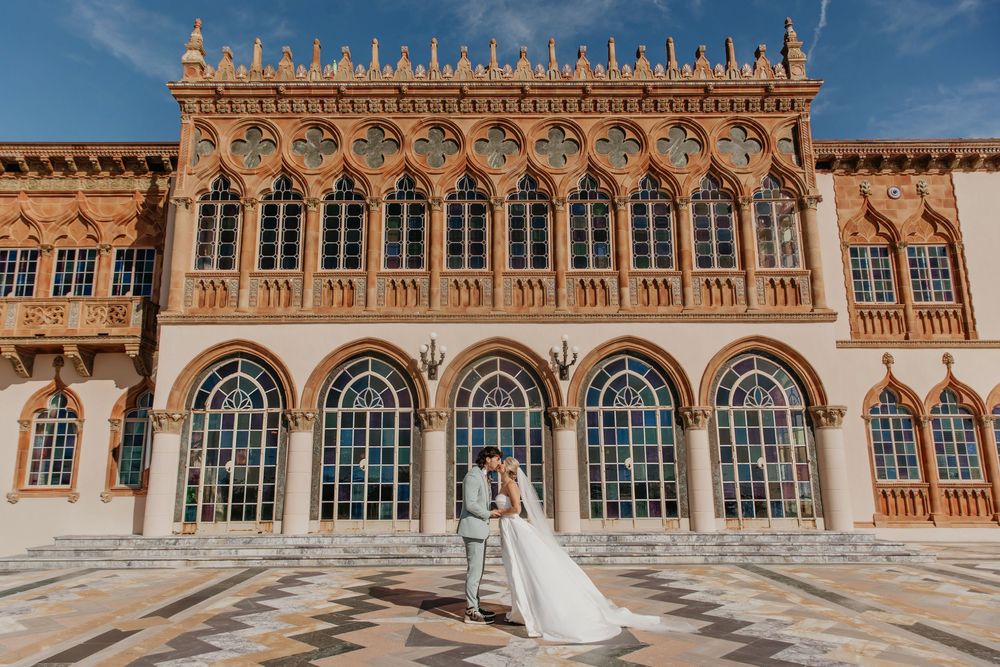 Bride in white wedding dress standing in front of ornate historic building with arched windows and decorative architecture.