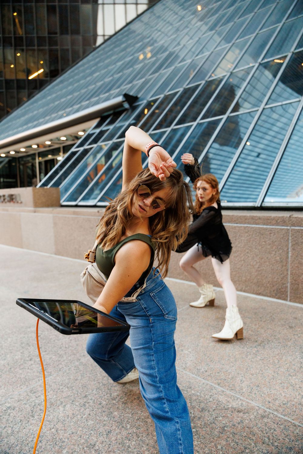 Two dancers posing outdoors near Houston's modern glass building, one holding a tablet with an orange cable.