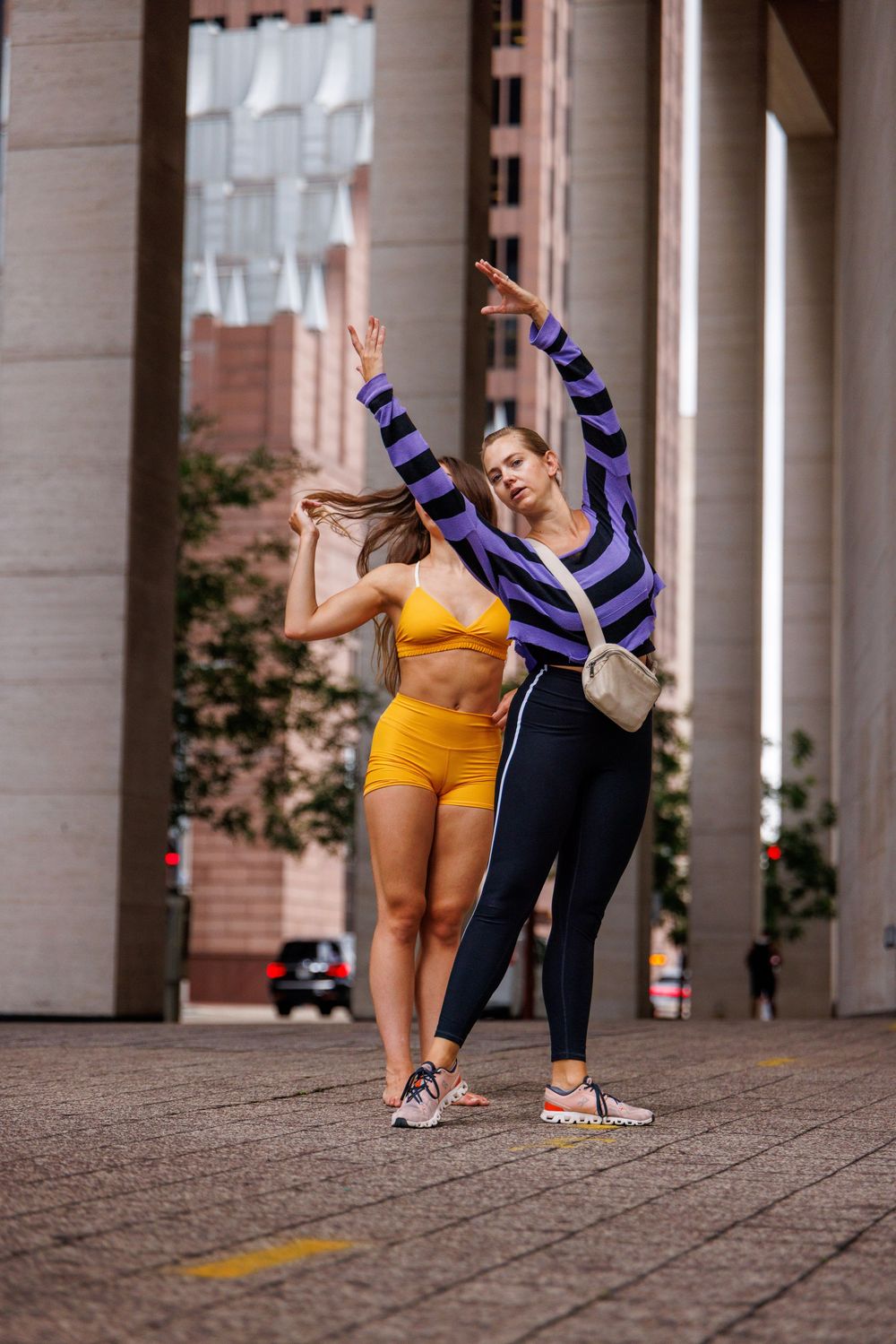 Two women dancing outdoors in urban setting, one in yellow sports bra and shorts, other in striped top and black leggings.