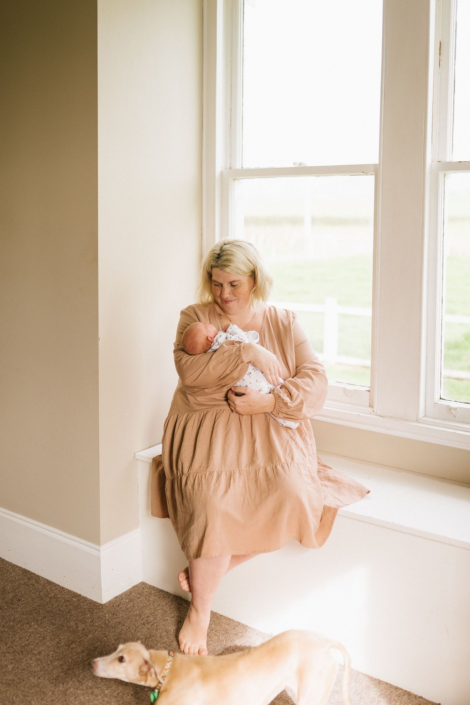 Mother in pink dress nursing baby by window with dog resting nearby.