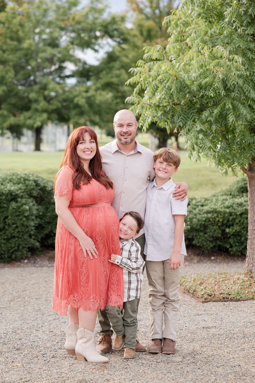 A family stands together outdoors, with a coral dress and casual attire against greenery and shrubs in the background.