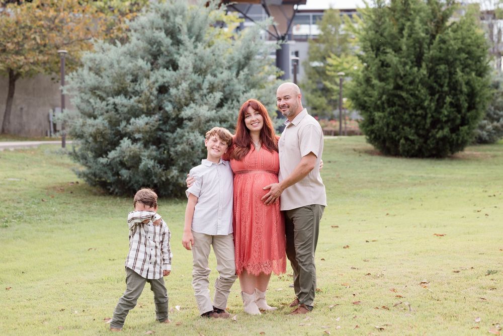A family group poses together on grass with evergreen shrubs and trees in the background.