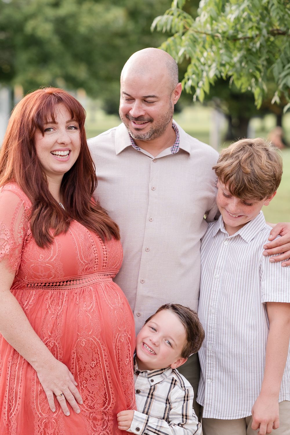 A family of four poses outdoors, with a pregnant woman in a coral dress and trees visible in the background.