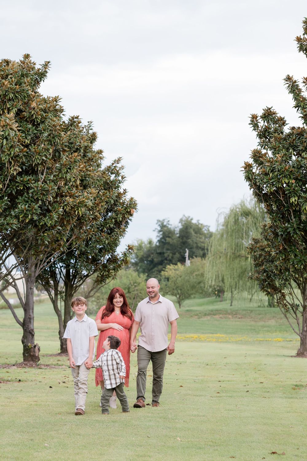 A family strolls through a park setting framed by large trees on a cloudy day.