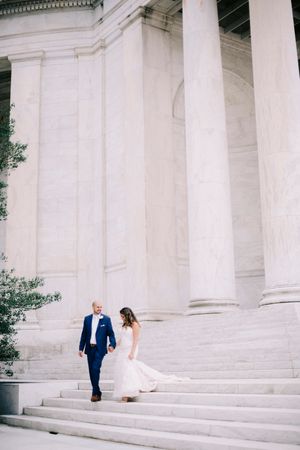 A couple in wedding attire poses on grand white marble steps of a classical building with columns.