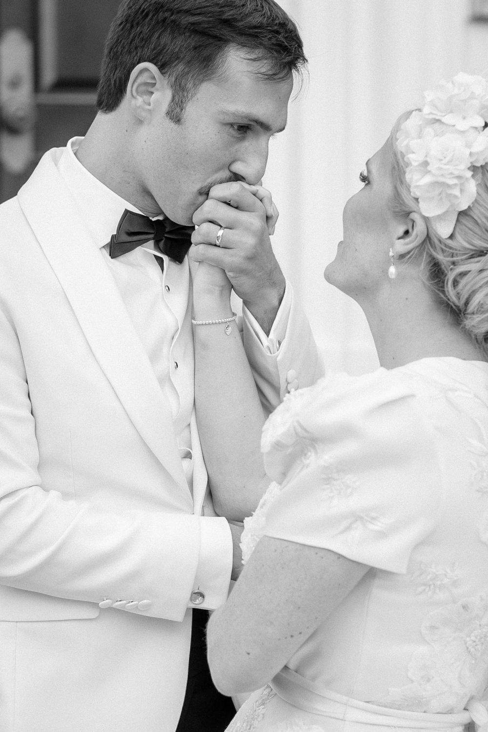 A tender black and white wedding moment shows a groom in white tuxedo kissing his bride's hand during their ceremony.