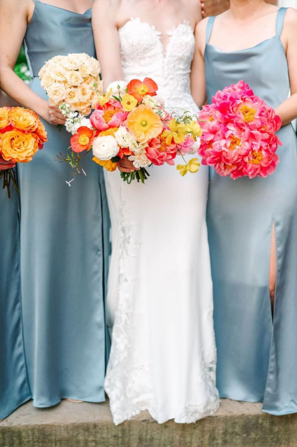 Bridesmaids in soft blue dresses hold vibrant coral and pink floral bouquets at a wedding ceremony.