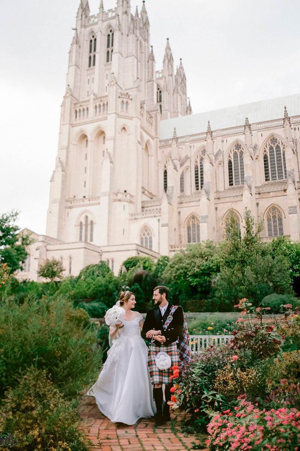 A romantic couple poses for wedding photos in front of the Gothic spires of the Washington National Cathedral with blooming roses.