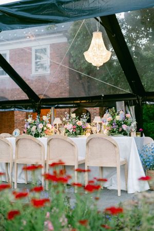 Elegant outdoor wedding reception under clear tent with chandelier, cane chairs, floral centerpieces, and red poppies in foreground.