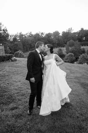 Black and white photo of a bride and groom sharing a kiss outdoors on a grassy lawn at dusk.
