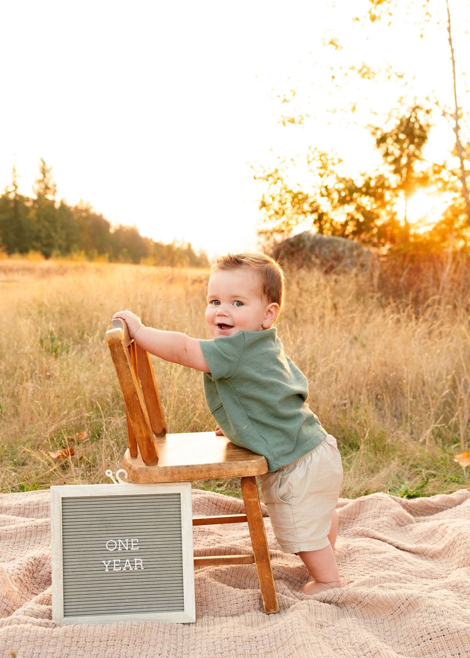 One year old toddler standing by wooden chair with letterboard sign during golden hour photoshoot.