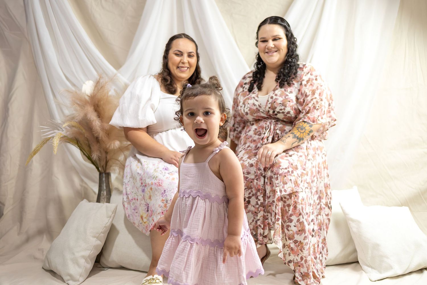 Two mums smiling behind an excited toddler girl in pink dress against white draped backdrop.