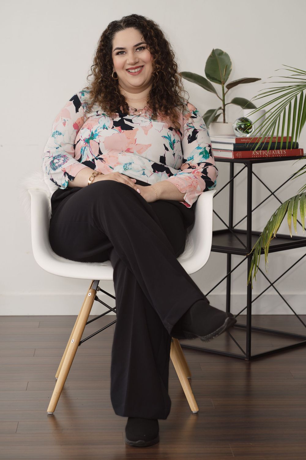 Relaxed business portrait sitting in white chair wearing floral top and black pants.