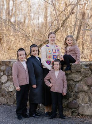 Five siblings posing together near a stone wall outdoors, one girl in a colorful crochet sweater.