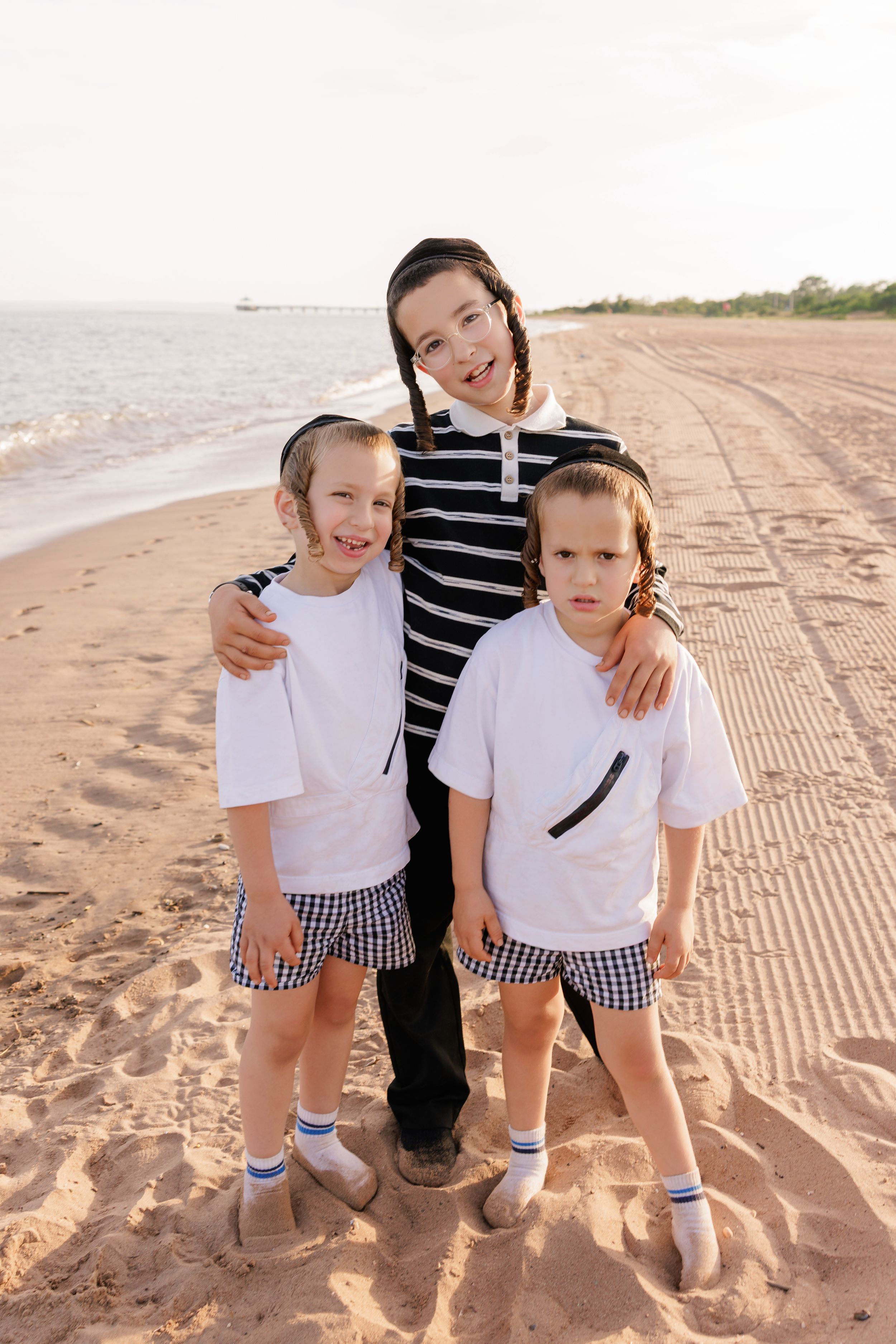Three children posing together on a sandy beach, two wearing matching white shirts and checkered shorts.