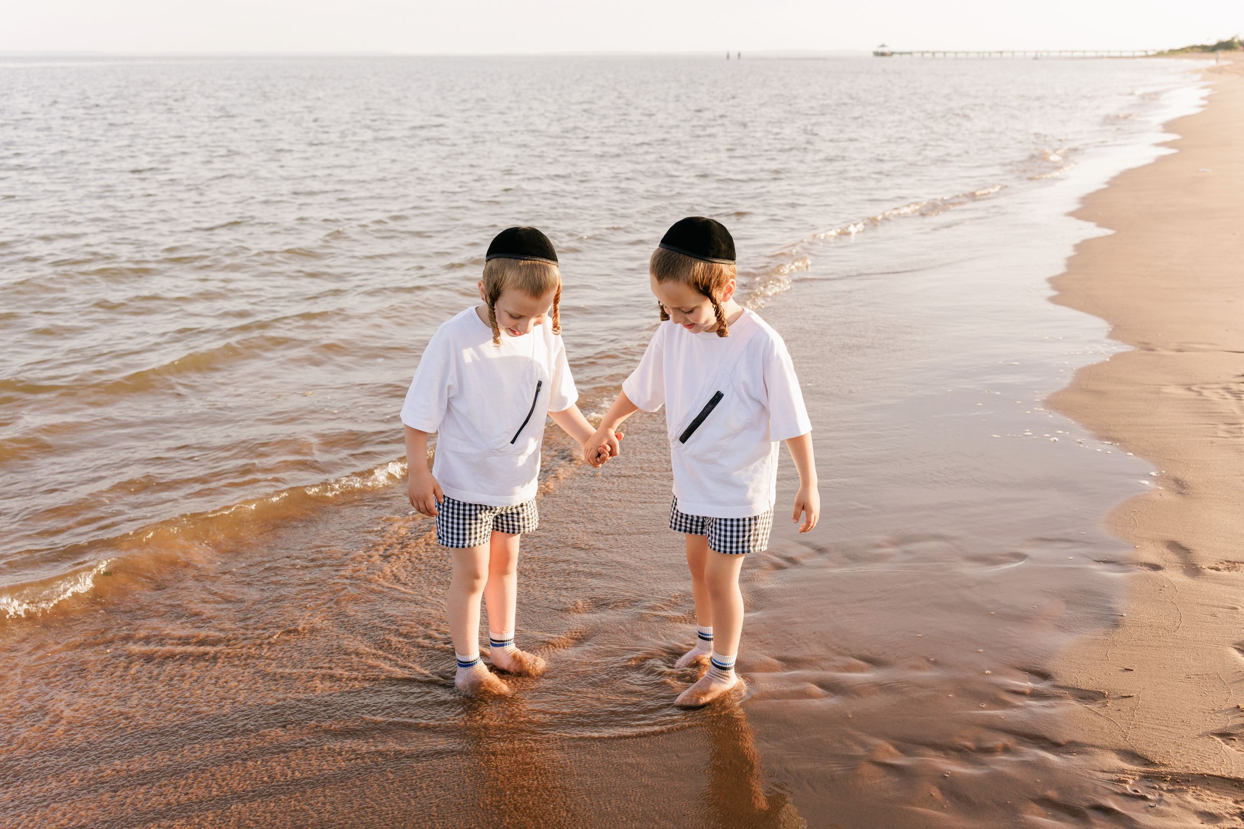 Two young children holding hands and walking along a sandy beach shore at sunset wearing white t-shirts.