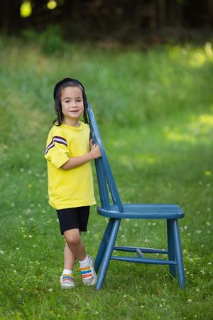 Young child in yellow shirt standing beside blue wooden chair on green lawn.