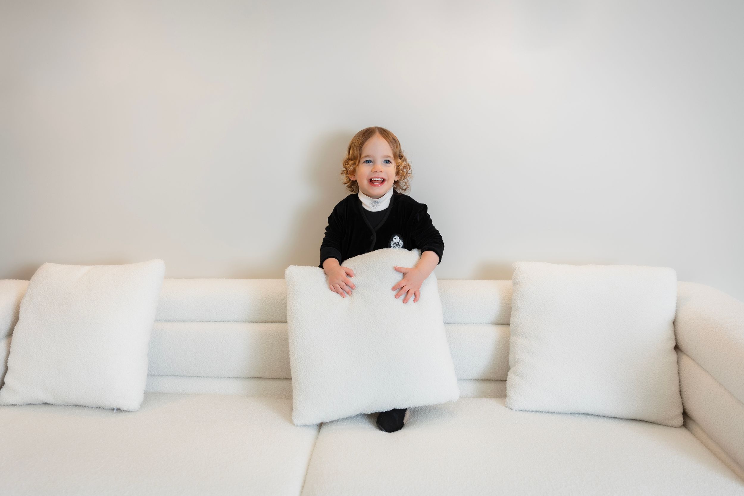 A happy toddler girl in a black dress holds a white pillow on a cream sofa against a light gray wall.