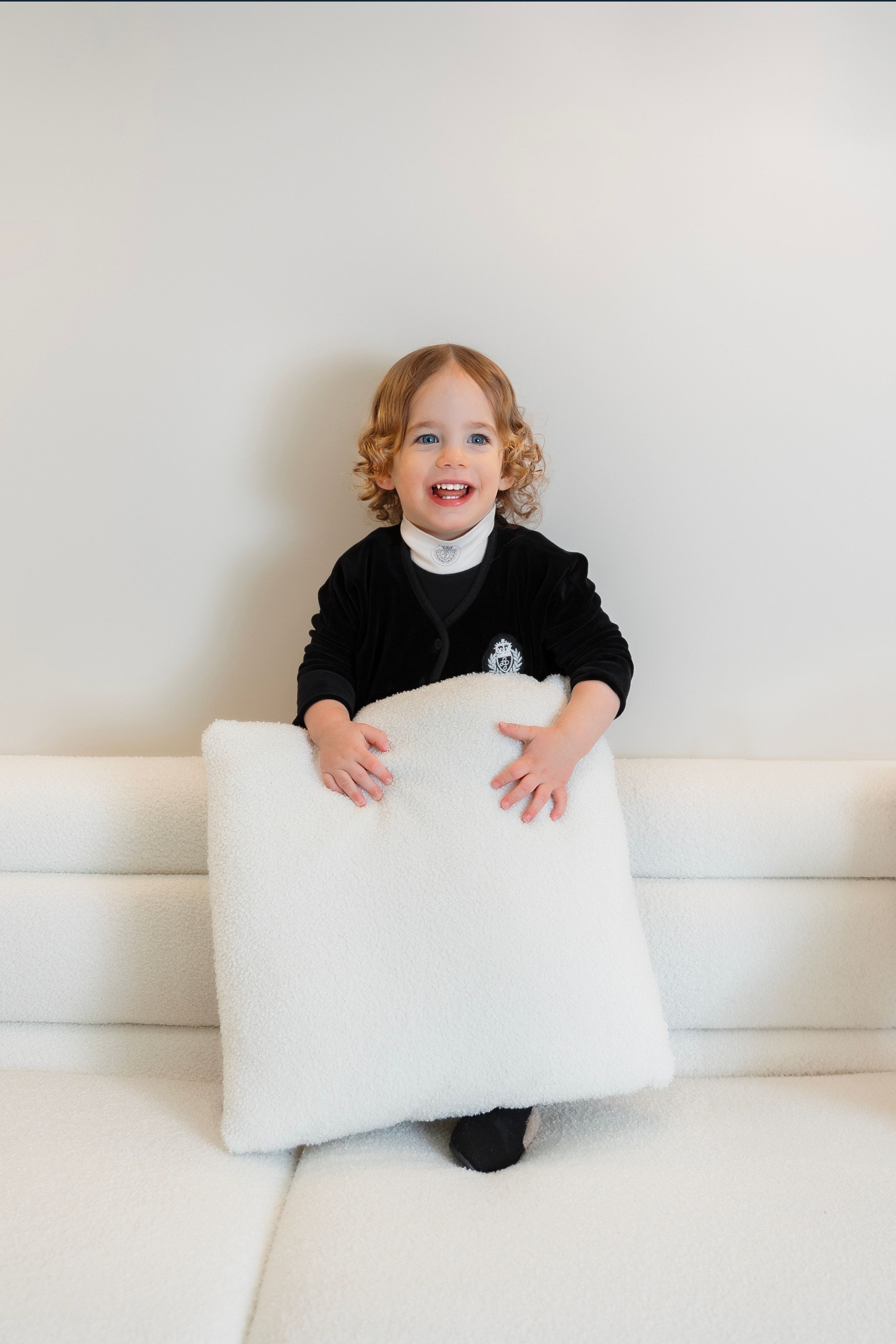 Smiling toddler in black outfit holding a white fluffy pillow while sitting on a white sofa.