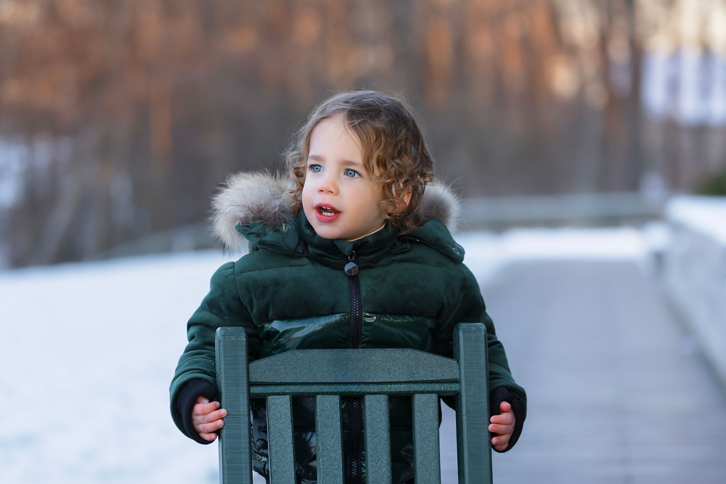 Toddler in green fur-hooded winter jacket sitting on a bench in a snowy park.