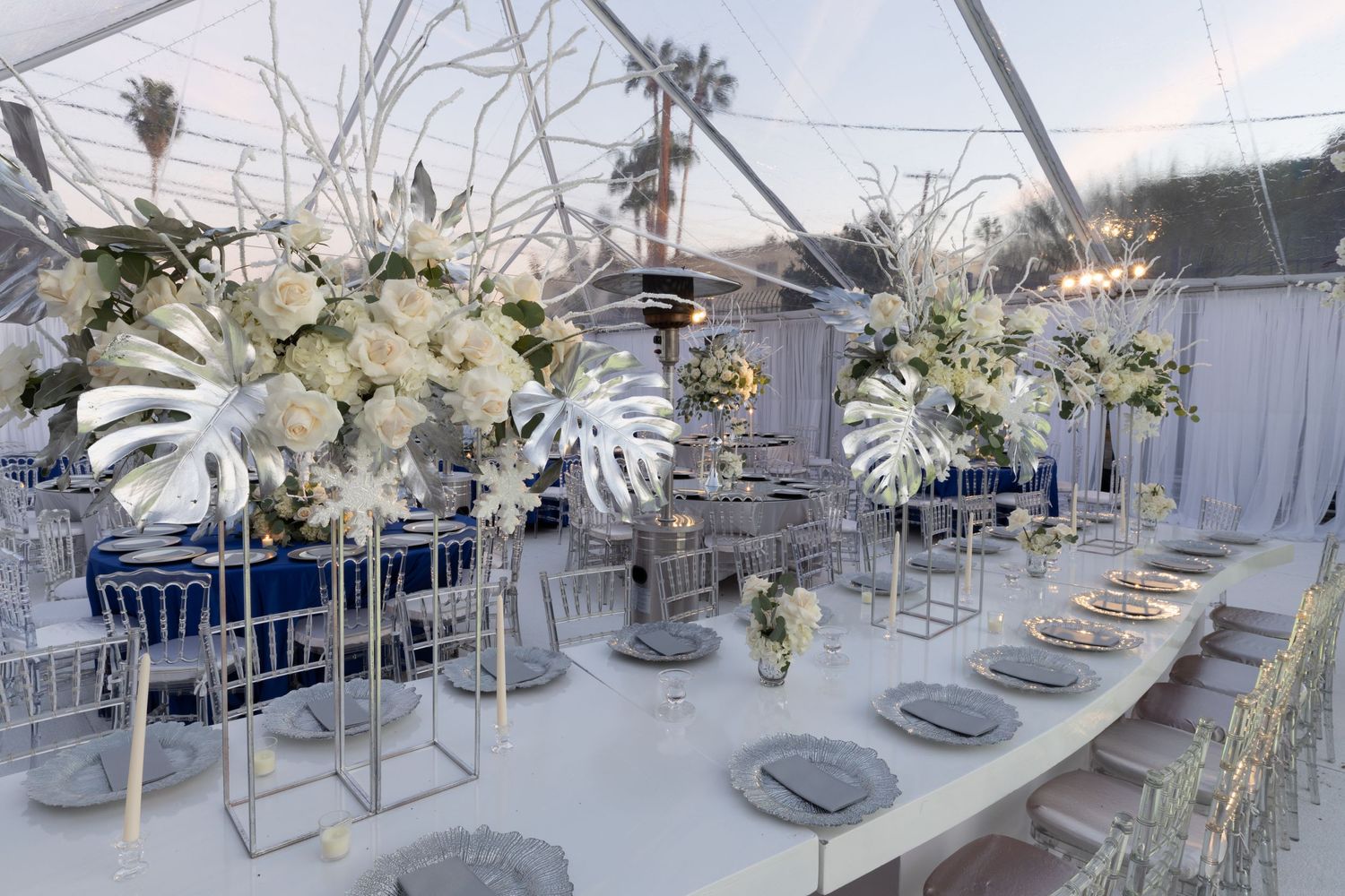 Elegant wedding reception table setting with tall white floral centerpieces and silver accents under a tent.