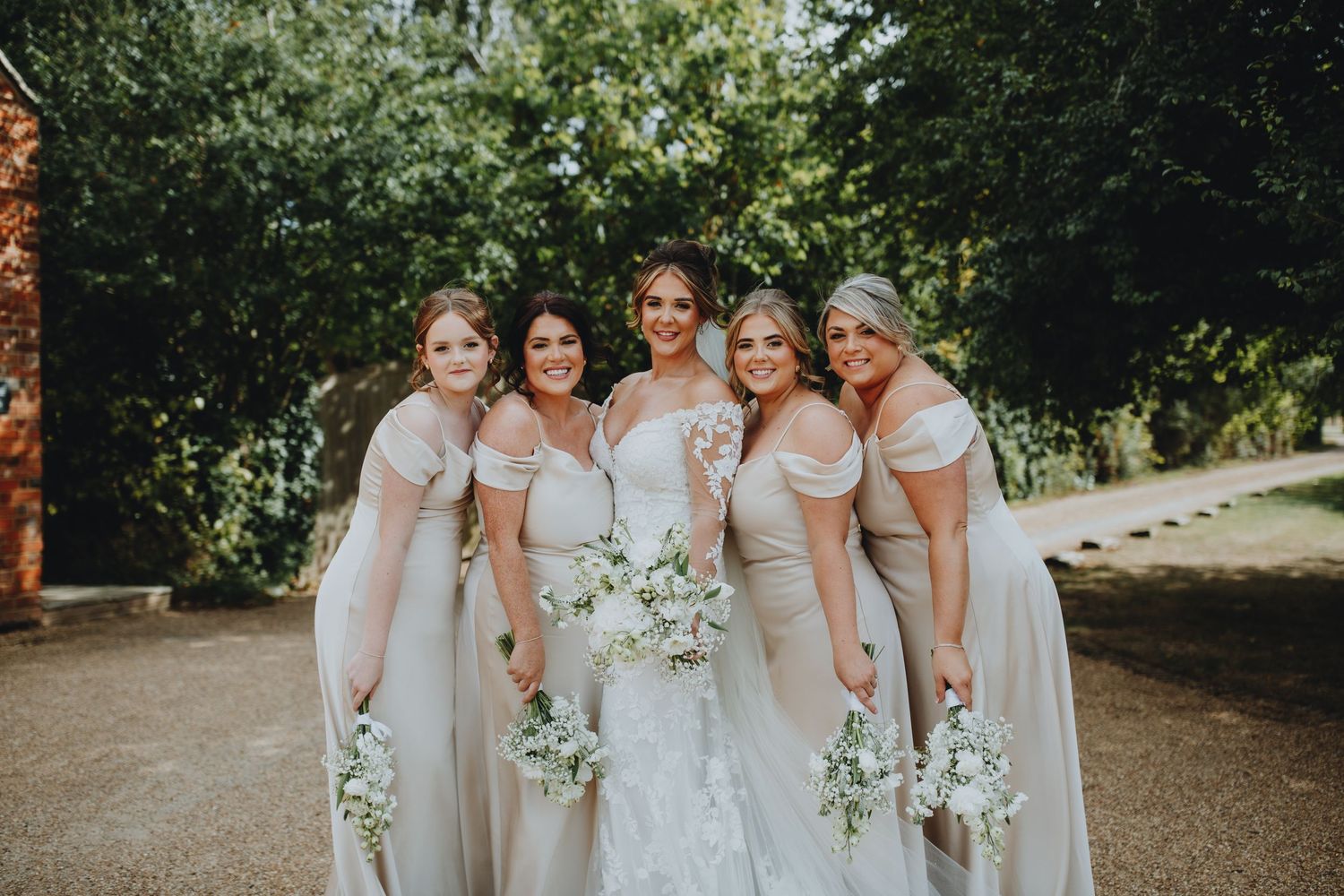 Wedding party in blush pink bridesmaid dresses and white bouquets pose together on path surrounded by greenery. Apton hall
