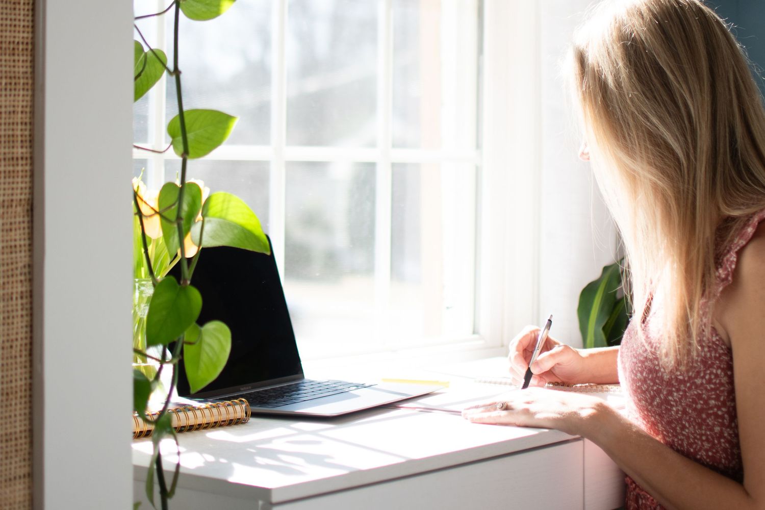 Woman writing at desk near window with houseplants and laptop in natural light.