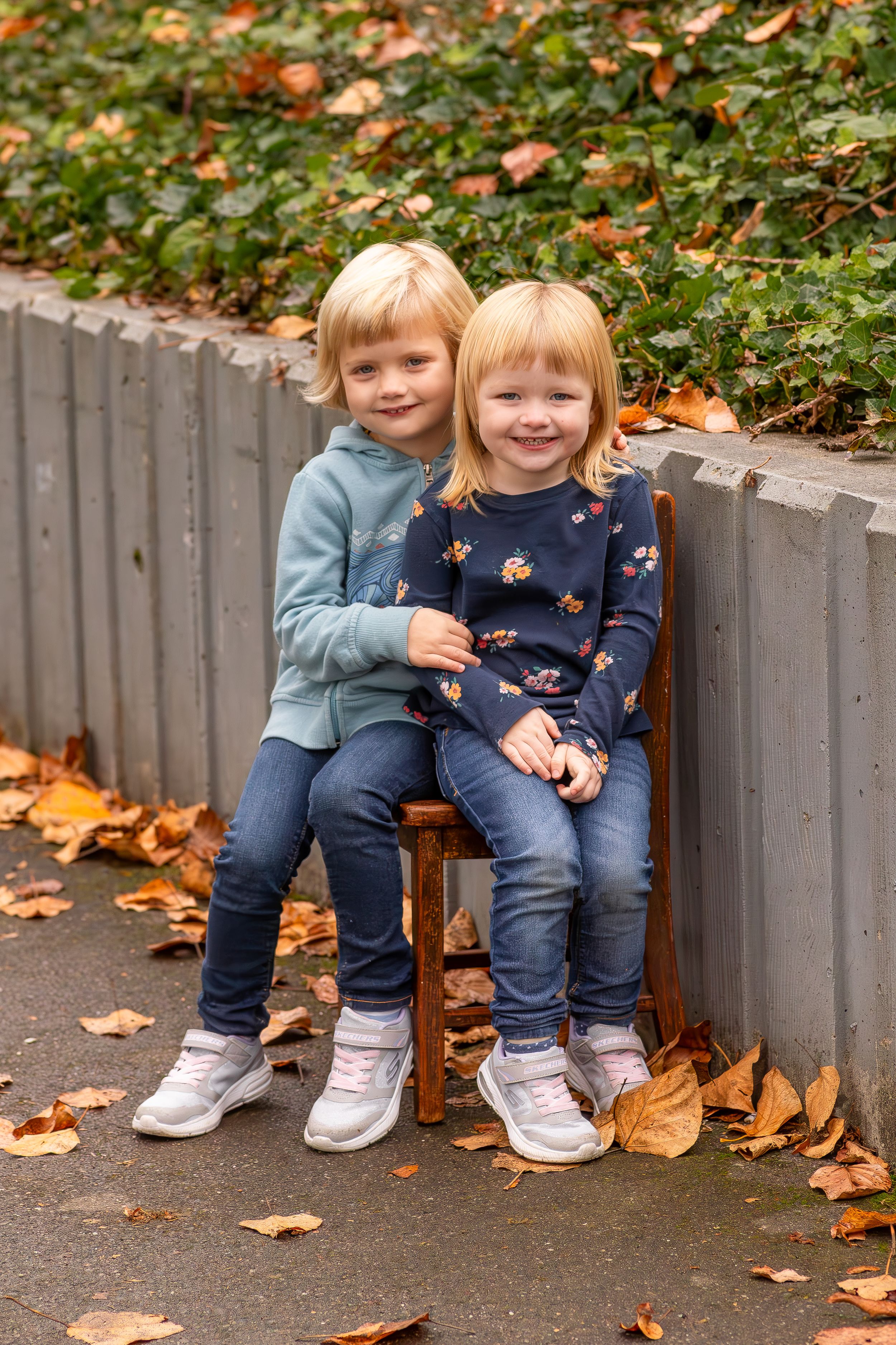Two small children in casual wear sitting on a wooden bench surrounded by autumn leaves.