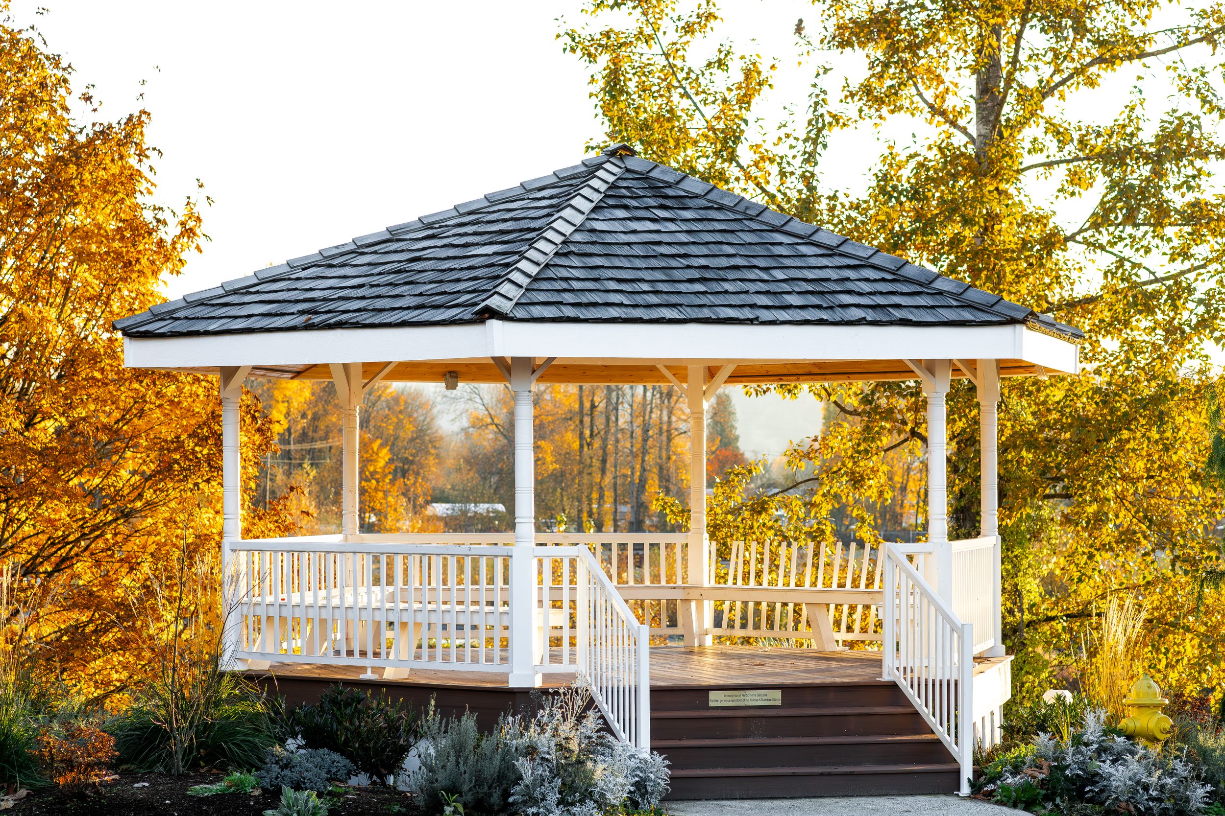 White gazebo with dark roof and wooden railings surrounded by autumn foliage.