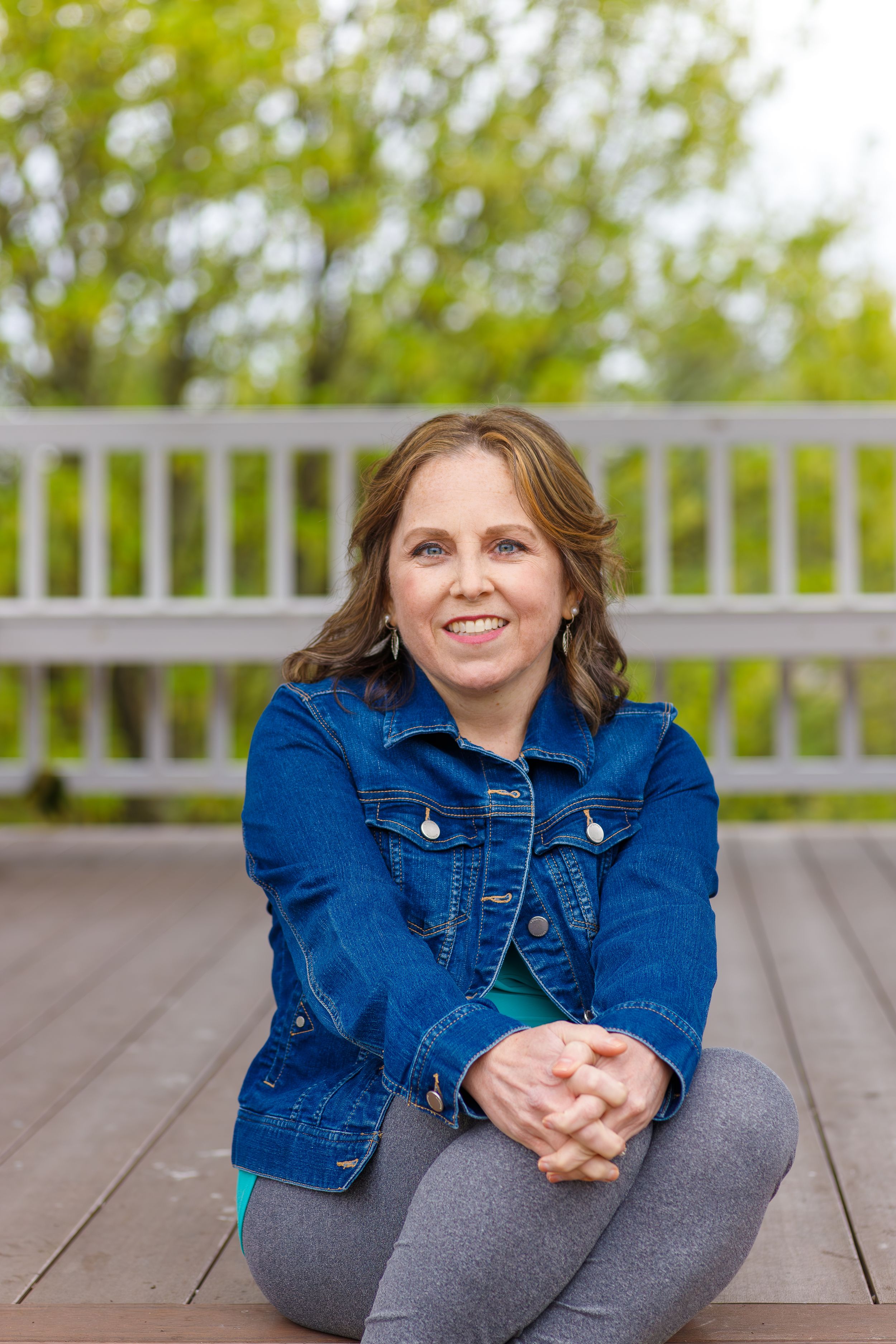 Woman in denim jacket sitting casually on wooden deck outdoors.