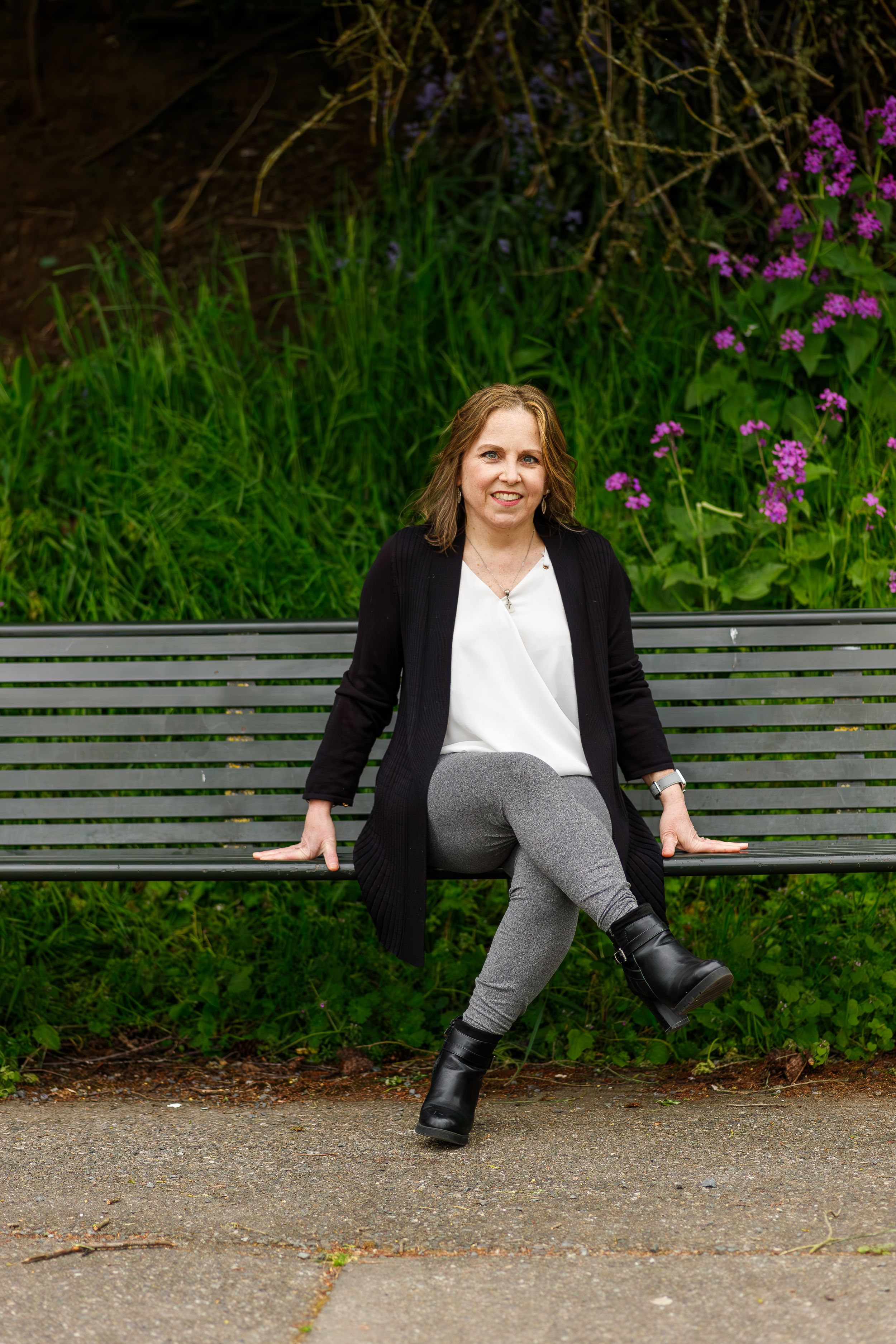 Woman in black cardigan and gray pants relaxing on park bench with flowering plants.