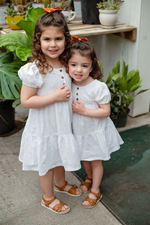 Two young siblings in matching white dresses and sandals pose together among potted plants.