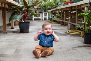 A baby in denim and khakis sits on concrete floor surrounded by potted plants.